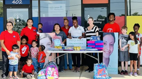 group of children of various ages are gathered around a table where merry maids is giving away free school supplies